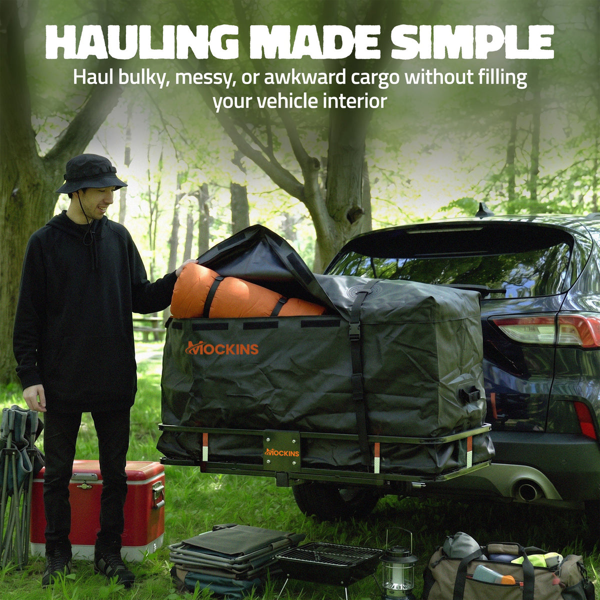 A person in black clothing stands beside a vehicle in a wooded outdoor setting, handling a large black Mockins cargo bag mounted on a trailer. The scene includes camping gear on the ground and a red cooler, with large promotional text at the top reading 'HAULING MADE SIMPLE.
