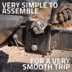 A gloved hand uses an adjustable wrench to tighten a bolt on a black hitch receiver labeled 'SMOOKINS'. Large white text reads 'VERY SIMPLE TO ASSEMBLE' at the top and 'FOR A VERY SMOOTH TRIP' at the bottom, with an outdoor gravel background.