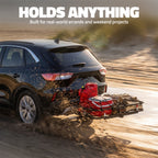 A black SUV driving on a dirt road while towing a large black trailer loaded with a red fuel canister and other gear. Large white text at the top reads 'HOLDS ANYTHING' with smaller text below, 'Built for real-world errands and weekend projects.